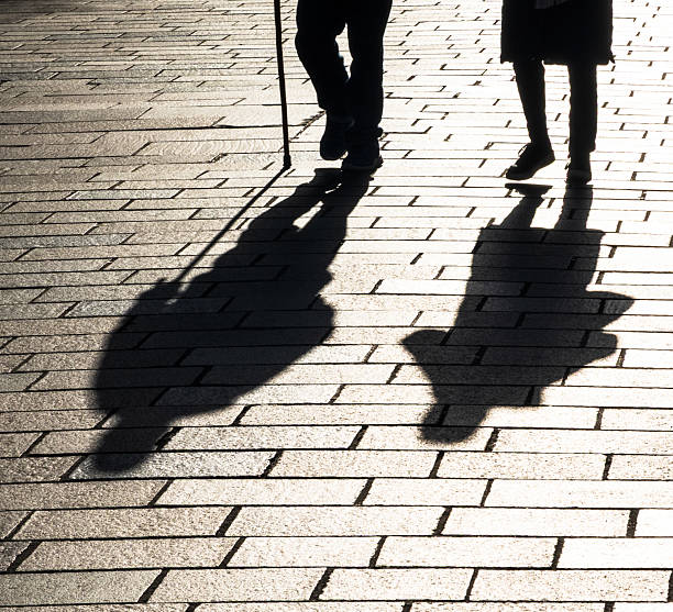 Shadows cast during winter, of an elderly man with a stick walks along the street with his partner.