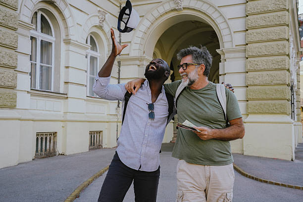 Two cheerful men, one Black and one White, walking together in an urban setting. They chat, laugh, and take selfies with a smartphone. Casual, diverse friendship portrayed in a warm, outdoor city environment. Perfect for themes like tourism, student life, modern friendship, or multicultural connection.