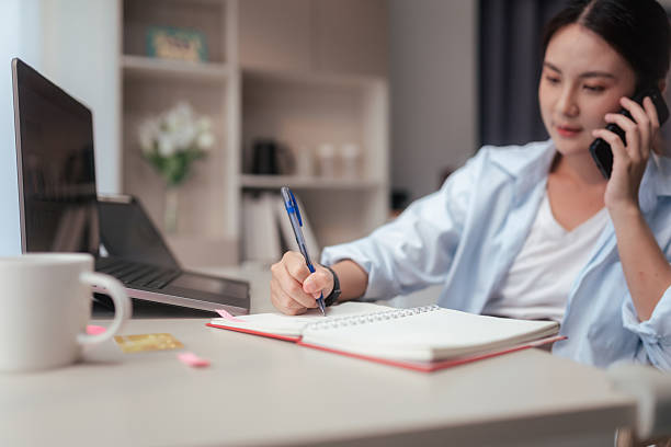 istock Young Asian woman multitasking while working from home, talking on the phone and taking notes in a notebook. Remote work, productivity, and modern freelance lifestyle in a home office environment. 2228956538