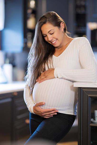 istock Pregnant woman smiling happily while embracing belly in a modern kitchen. 2228962415