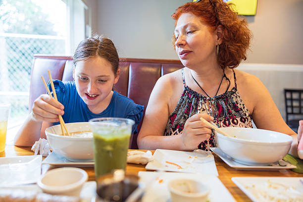 istock Smiling girl learns eating with chopstics technique from 47-year-old mom. Authentic Asian cuisine experience in bright restaurant booth. 2228963397