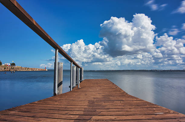 Scenic view of a wooden pier extending over calm coastal waters under a vivid blue sky with large, dramatic clouds. The perspective draws the viewer toward the horizon, evoking a sense of peace, freedom, and connection to nature. Ideal for themes such as travel, relaxation, nature, serenity, and coastal lifestyle. Perfect for use in tourism materials, blogs, or meditation and wellness visuals.