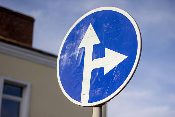 This image showcases a blue round road sign featuring white arrows pointing straight ahead and to the right. The sign is positioned near a building and against a sky background, indicating directions at a crossroads. It is suitable for themes related to navigation, orientation, or traffic control.