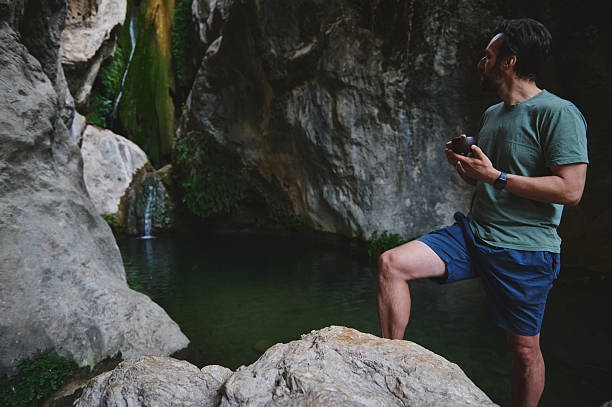 istock Man Relaxing by Serene Waterfall Amidst Rocky Natural Scenery Holding a Cup 2228994243