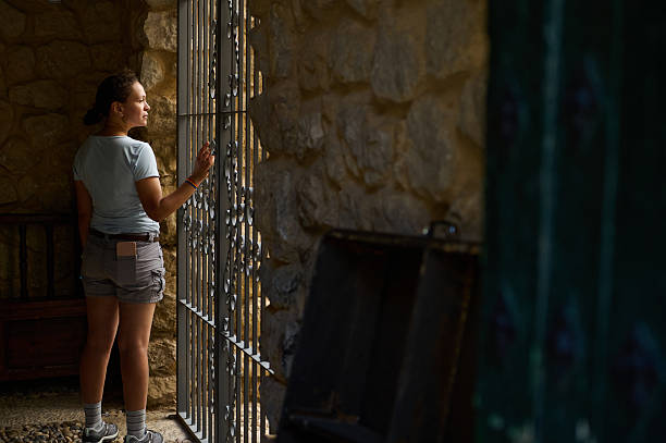 A woman, standing by a weathered wall, gazes outward through a decorative iron window, surrounded by warm, rustic tones and shadows, suggesting a tranquil and reflective ambiance.
