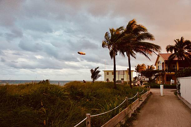 A dramatic north-facing view along the Miami Beach coastline captures windswept palm trees and golden storm clouds at dusk, as an iconic blimp floats above the Atlantic Ocean. Typical pastel houses and lush dune grass evoke Florida’s relaxed beach lifestyle, while the Goodyear Blimp—frequently seen along Miami’s coast for major events—adds a touch of vintage aviation nostalgia. The scene perfectly illustrates Miami’s blend of tropical energy, unpredictable weather, and legendary coastal imagery.
