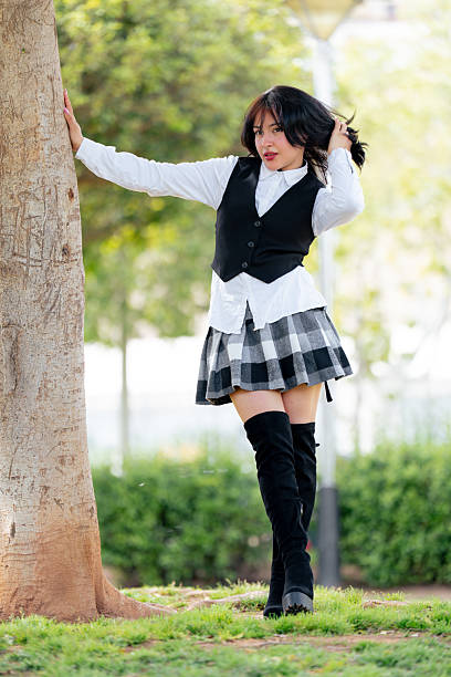 Fashionable young woman posing in a sunlit park. Leaning against a tree while playfully touching her hair. Exuding confidence and happiness in a stylish outfit of a white shirt. Black vest. And skirt