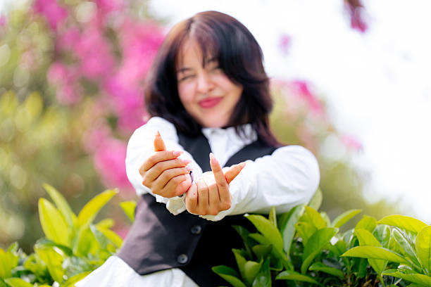 Young woman wearing a white shirt and black vest making a money gesture with her hands, standing in front of a blooming tree adorned with vibrant pink flowers