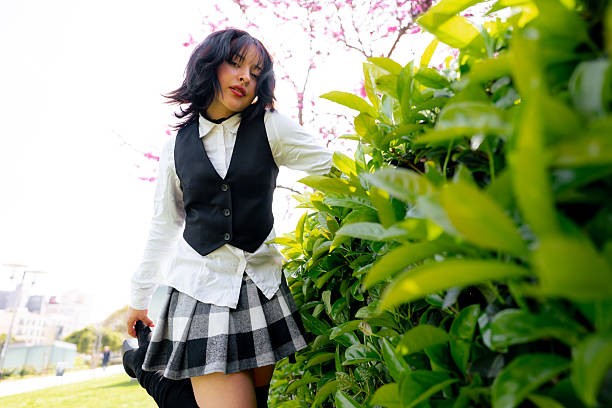 Low angle view of a young student posing in a park near a hedge and a flowering tree, wearing a white shirt, black vest and a checkered skirt