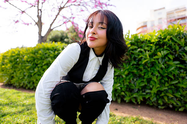 Young woman with nose piercing and modern clothes crouching in a park looking at camera on a sunny day with a flowering tree in the background