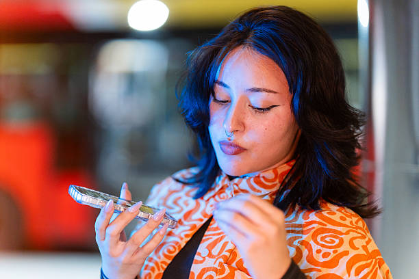 Young woman retouching her make-up with a smartphone as a mirror, standing in a vibrant public space illuminated by neon lights, exuding elegance and concentration