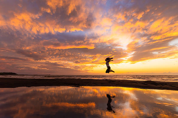 A woman doing a jump with knees tucked and side view with epic sunset and tide pool reflections.