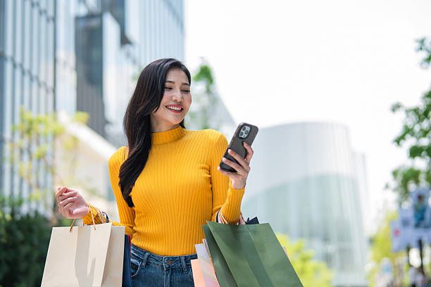 Asian young woman using mobile phone while stand in department store. Attractive beautiful female holding shopping bags walking outdoors with happiness enjoy purchasing in shopping mall center.