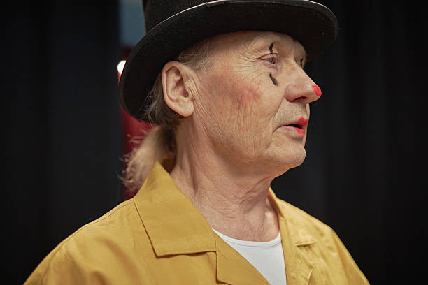 Senior Caucasian man wearing clown makeup and black top hat standing in profile during talent competition, visible wrinkles and painted red nose emphasizing expressive character
