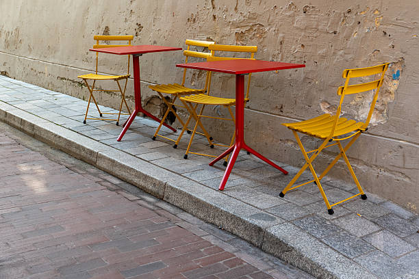 A charming scene of outdoor cafe seating in Vilnius Old Town, Lithuania, during sunset. Yellow chairs and red tables are set along a historic, cobblestone street with rustic buildings in the background. The warm, golden evening light highlights the historic architecture and lively street atmosphere, ideal for travel, lifestyle, and European cityscape themes.