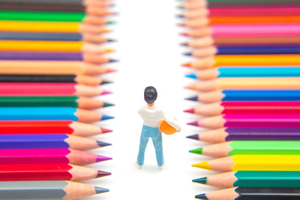 A small worker figurine holding a tool navigates between rows of vivid colored pencils, symbolizing creativity in labor on a minimalist white background