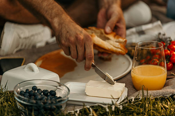 Hands spreading butter on a slice of bread during a picnic on a blanket on grass, with blueberries, cherry tomatoes, and orange juice