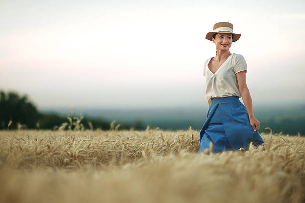 Young woman wearing a stylish hat walking through a golden wheat field, smiling joyfully as the sun sets, embracing the beauty of nature