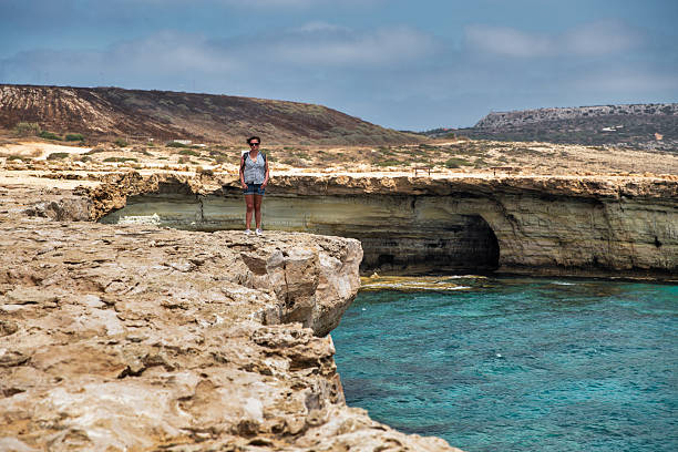 Seascape Cape Greco peninsula park with woman standing over a cliff, Cyprus. It is mountainous peninsula with a national park, rock paths, a turquoise lagoon and a natural stone bridge.