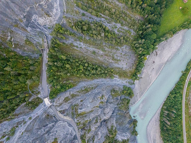 Drone shot of winding mountain road crossing rocky terrain near turquoise river. Scenic landscape with forested hills and natural river curves. Mountainous outdoor environment with road infrastructure.