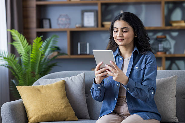 istock Young Indian girl sitting on sofa at home, using tablet, talking on video call 2229038926