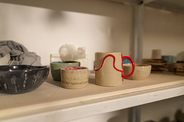 Various handmade clay mugs and bowls with unique designs are drying on a shelf in a pottery workshop, showcasing the creativity and craftsmanship of the ceramic artist