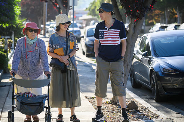Elderly woman walking using a mobility walker on the pedestrian footpath, accompanied by her adult children. California.