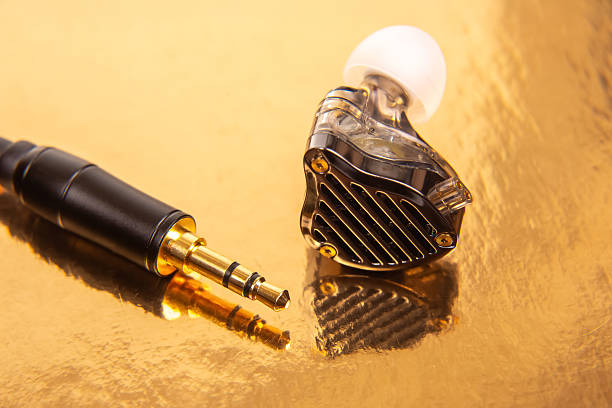 istock A single earbud, a gold-plated 3.5mm jack, and a black audio device on a reflective golden surface, ideal for tech product shots and audio themes. 2229052043