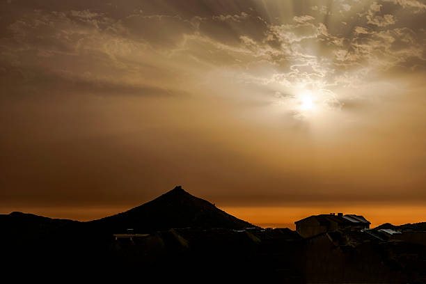 Yellow and orange hues in a cloudy sky. Radiant sunbeams pierce through the clouds above a mountain, casting light over rooftops in backlit shadow.