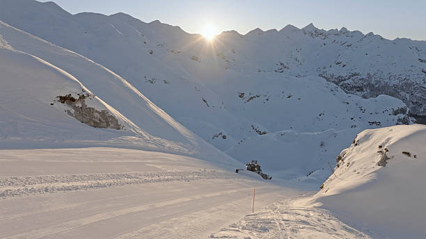 Snow-covered mountain ski slope with freshly groomed tracks under a bright sunrise. The serene alpine landscape features rugged peaks and soft white snow.
