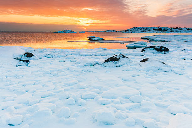 Snow blankets the coastal landscape as the sun sets in Sweden, casting warm colors across the icy waters and highlighting the rocky shoreline. Tranquility fills the air at dusk.