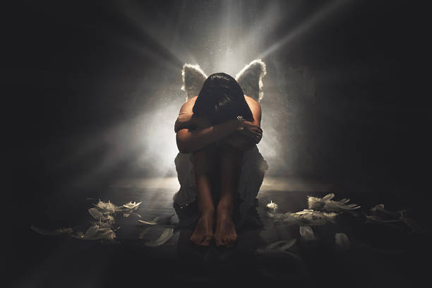 A young man dressed in a fallen angel costume with black wings looks sullenly at the camera. Girl white angel standing behind dark angel. Shooting on the roof at night