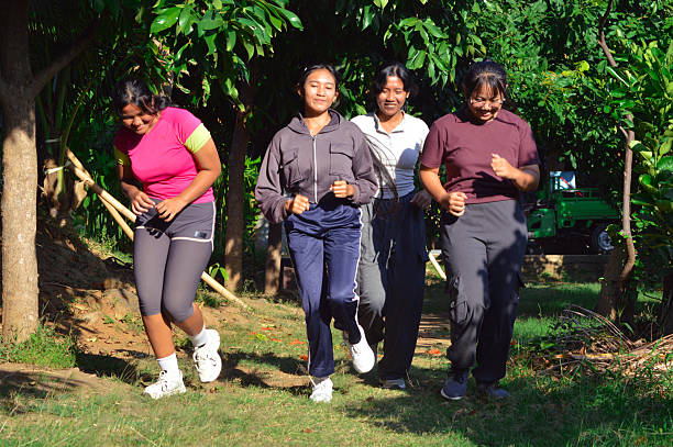 Four teenage girls continue jogging together under tropical trees, playfully ducking and dodging branches while laughing in the sunny garden.