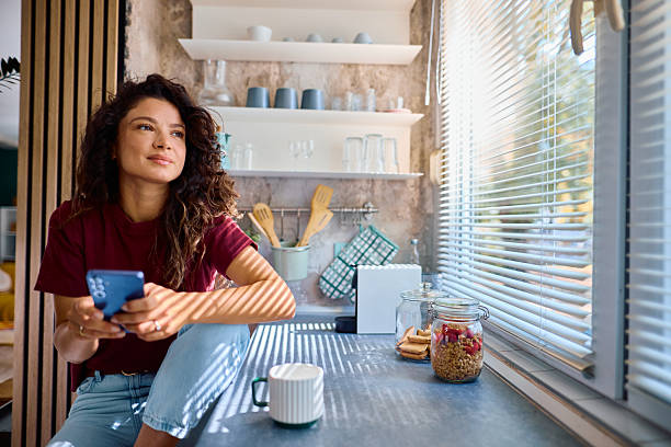 istock Young woman holding smartphone and looking away while sitting in kitchen 2229088572