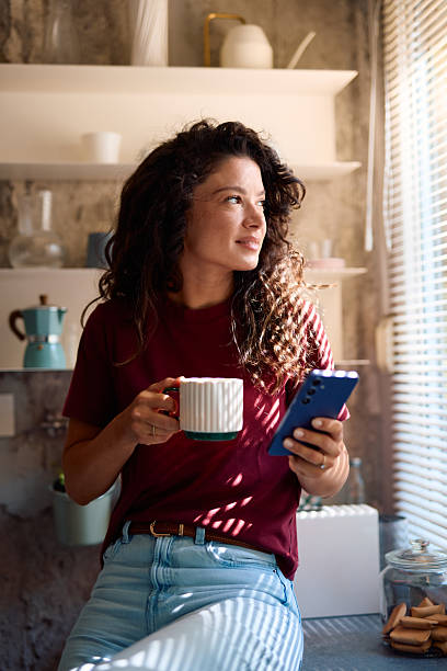 istock Young woman enjoying a cup of coffee and using smartphone in kitchen 2229088597