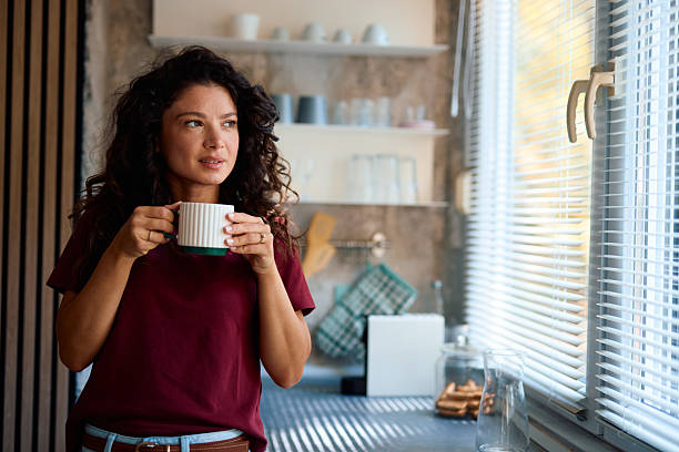 istock Young woman drinking coffee looking out the window in her kitchen 2229088668