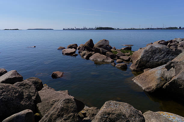 Seaside landscape in Kotka, Finland with a rocky shoreline, calm waters, and a distant industrial port on a clear summer day.