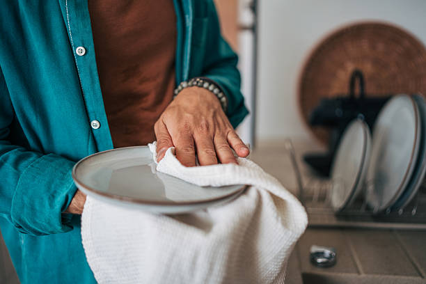 Man is carefully drying a plate with a white dishcloth after washing it, standing in a modern kitchen with other clean dishes in the background