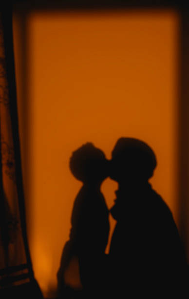 A tender, softly blurred silhouette captures a mother, wearing a hijab, leaning in to lovingly kiss her child. The moment is set against a warm, orange background, creating an intimate and emotional scene that symbolizes the deep love, security, and profound bond between a parent and their child.