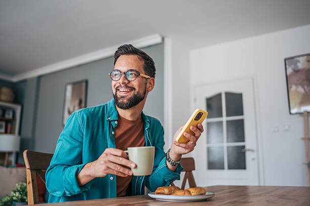Happy man sitting at a dining table, enjoying breakfast with a warm cup of coffee in hand while browsing his smartphone, smiling and soaking in the cozy atmosphere of home