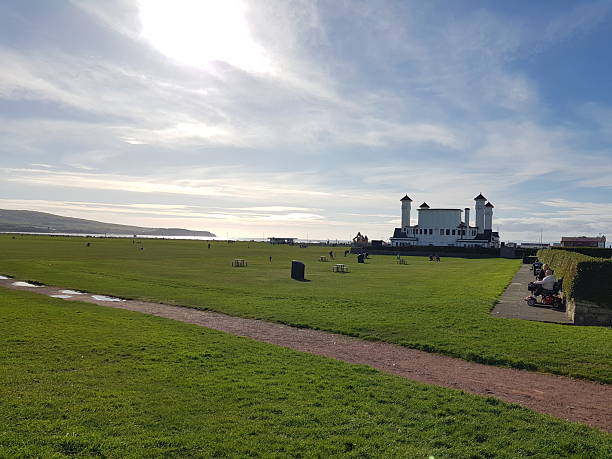 Ayr, Scotland, UK - March 24, 2024: Low Green, grass area overlooking Ayr Bay in South Ayrshire. People enjoying a sunny day at the grassland. At the background amusement centre