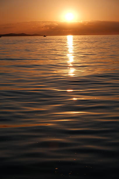 Stunning sunset over the Adriatic Sea captured from the coastline of Zadar, Croatia. The sky glows with vibrant hues of orange, pink, and purple, reflecting beautifully on the calm water. Silhouettes of ancient architecture, boats, and the famous Zadar Sea Organ add depth and character to the scenic view. Perfect for travel, nature, landscape, and European destination concepts.