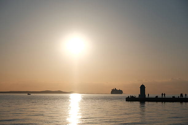 Stunning sunset over the Adriatic Sea captured from the coastline of Zadar, Croatia. The sky glows with vibrant hues of orange, pink, and purple, reflecting beautifully on the calm water. Silhouettes of ancient architecture, boats, and the famous Zadar Sea Organ add depth and character to the scenic view. Perfect for travel, nature, landscape, and European destination concepts.
