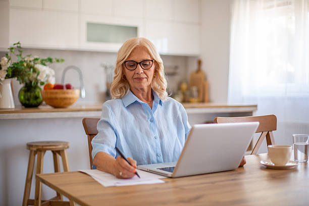 istock Senior adult woman working from home using laptop and taking notes 2229116613
