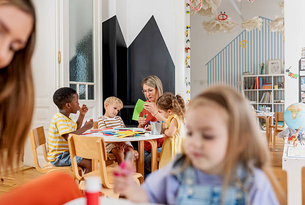 istock Two preschool teachers helping diverse children with arts and crafts using scissors, glue, and colorful paper in a cheerful classroom setting 2229128505