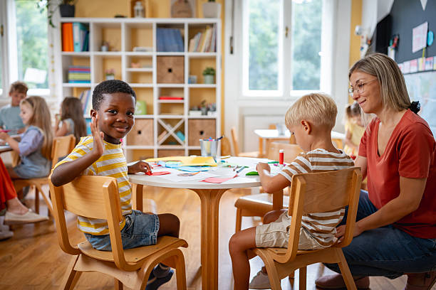 Smiling boy waving at the camera while sitting with classmates and teacher during a multicultural preschool craft activity