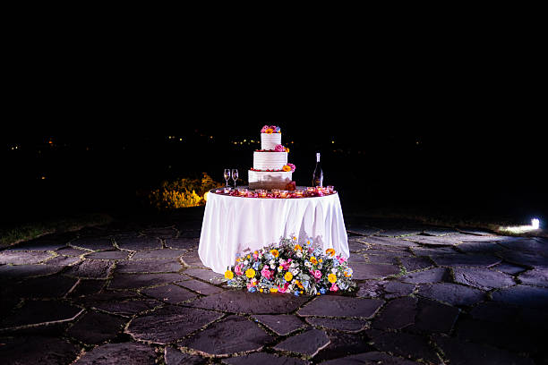 A beautifully arranged wedding cake on a table surrounded by flowers, set against a night sky with distant lights twinkling.