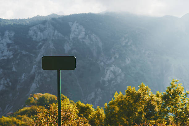 Dark-green rectangular signpost mock-up against scenic mountain landscape with lush green trees in sunlight and misty rocky peaks in background, perfect for template or design placement; a copy space