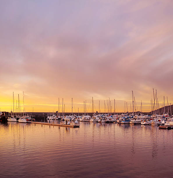 A tranquil marina scene illuminated by a breathtaking sunset, showcasing docked sailboats and yachts under a vivid, colorful sky reflecting on calm water. Perfect for conveying serenity, beauty, and peacefulness.