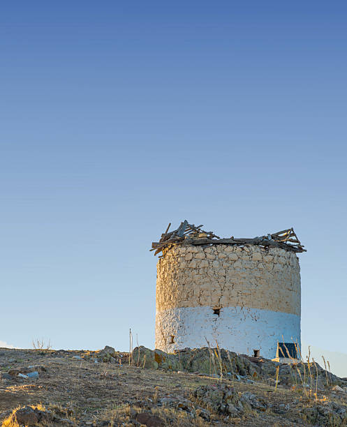 An old stone structure, weathered and partially ruined, rests prominently on an open hillside against a clear blue sky. Surrounded by sparse vegetation, it exudes history and solitude in a rural location.